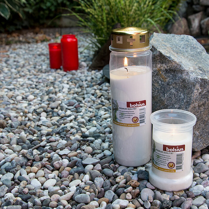 Im Bild sind zwei brennende Bolsius Grabkerzen zu sehen, eine größere und eine kleinere, die auf einem Kiesbett neben einem großen Stein platziert sind. Im Hintergrund sind unscharf zwei weitere rote Bolsius Grabkerzen zu erkennen. Die Szenerie ist in ein