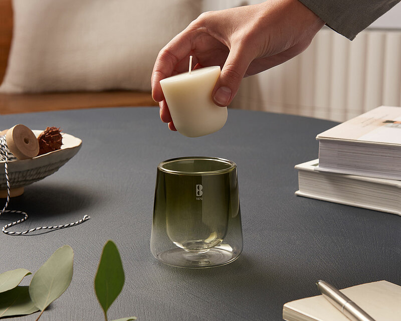 Es ist ein ruhiges und klares Bild mit aufeinander abgestimmten Farbtönen. In einem gemütlichen Wohnzimmer auf einem dunkelgrauen Wohnzimmertisch steht ein Bolsius Cleanlight Glas. Man sieht wie ein Mensch das Cleanlight Nachfüllwachs in dem Glas platzieren möchte. Auf dem Tisch befinden sich mehrere Bücher und ein paar grüne Zweige.