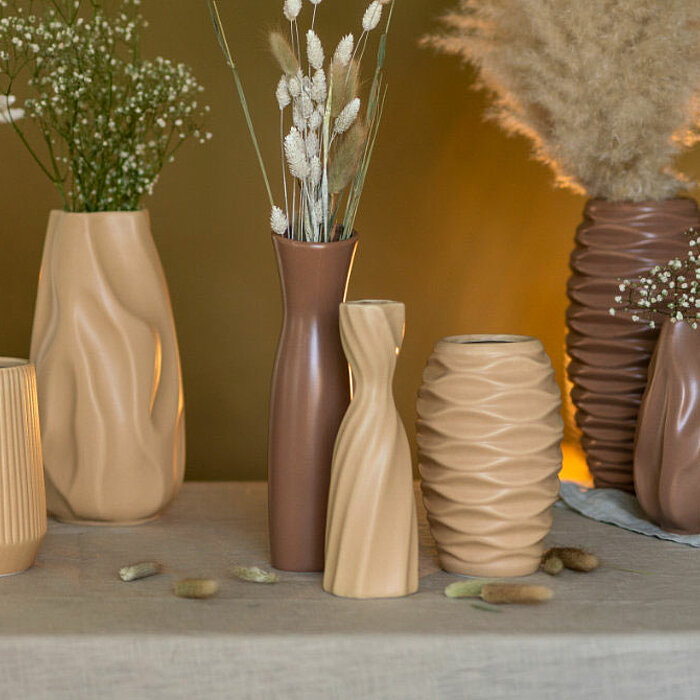 Acht Vasen vom Hersteller "Sandra Rich" in verschiedenen Formen und Erdtönen stehen auf einem beige gedeckten Tisch. Die Vasen enthalten diverse Trockenblumen wie Pampasgras und Schleierkraut. Auf dem Tisch sind kleine Steine und Stofftücher verteilt, was