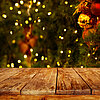 Ein rustikaler Holztisch steht im Vordergrund, während im Hintergrund ein verschwommenes, festlich dekoriertes Weihnachtsbaum zu sehen ist. Der Baum ist mit zahlreichen leuchtenden Lichtern, roten und goldenen Kugeln sowie anderen traditionellen Weihnacht