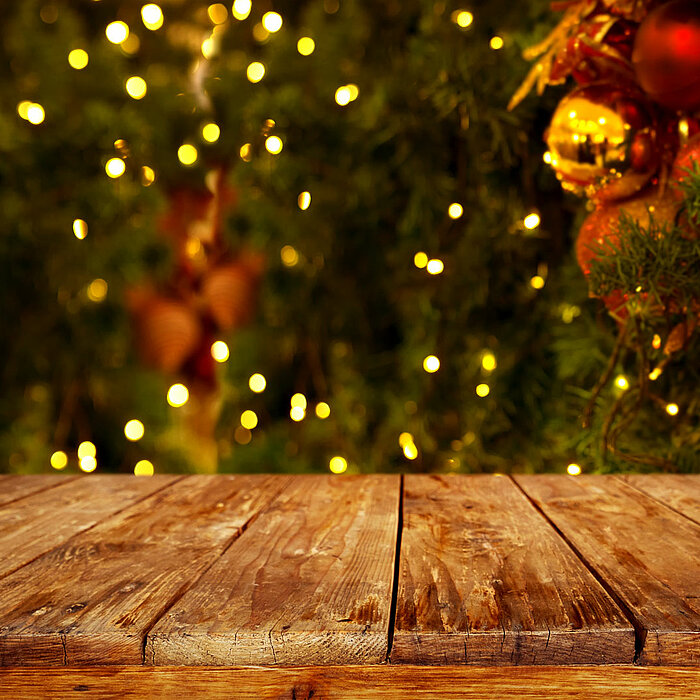 Ein rustikaler Holztisch steht im Vordergrund, während im Hintergrund ein verschwommenes, festlich dekoriertes Weihnachtsbaum zu sehen ist. Der Baum ist mit zahlreichen leuchtenden Lichtern, roten und goldenen Kugeln sowie anderen traditionellen Weihnacht