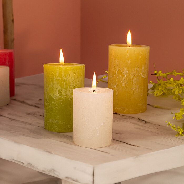 Drei brennende Bolsius Rustikkerzen in den Farben Magenta, Rot und Gelb stehen auf rustikalen Holzplatten. Umgeben sind sie von weißen und pinken Tulpen sowie kleinen grünen Zweigen. Der Hintergrund besteht aus hellen Holzbrettern, die eine warme und gemü
