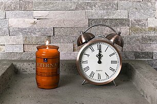 Auf dem Bild auf einem eckigen Betonuntergrund steht ein rotes brennendes Öllicht Nr. 3 von Aeterna und daneben ein schöner, klassischer kupferfarbener Wecker. Der Wecker hat eine traditionelle Form mit zwei Glocken und Zeigern auf einem weißen Zifferblatt. Im Hintergrund ist eine raue Steinmauer zu sehen, deren natürliche Textur und Farbe dem Bild eine rustikale und charmante Atmosphäre verleihen.