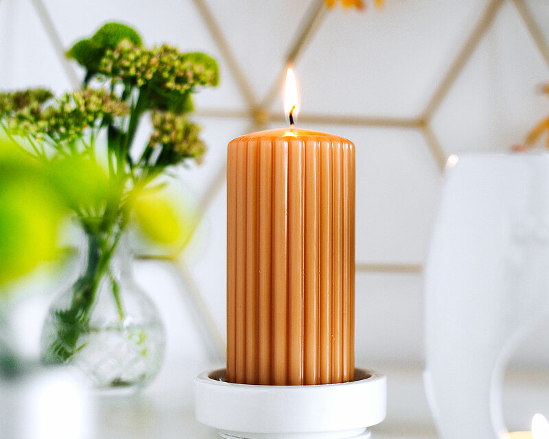 Das Bild ist in hellen und freundlichen Farben es strahlt Freundlichkeit und Entspannung aus. In der Mitte Des Bildes auf einem weissen Tisch steht ein weisser Kerzenhalter auf dem Kerzenhalter steht eine orangefarbene Duftstumpenkerze geriffelt von Bolsius. Am rechten Bildrand steht ein weisses Duftstövchen oval von Bolsius mit einem brennende Teelicht. Auf dem Tisch stehen verschiedene mit Wasser gefüllte Glasvasen in die Blumen gesteckt sind.