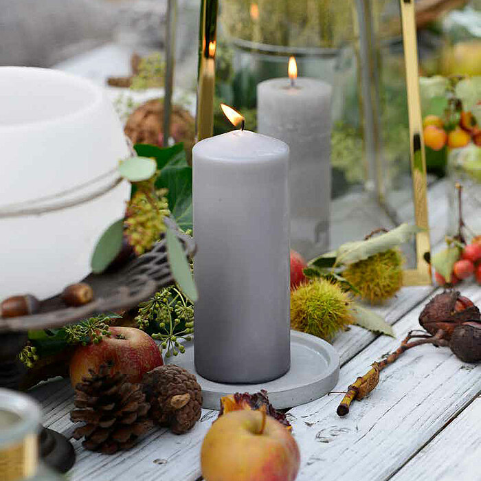 Eine brennende graue Stumpenkerze vom Hersteller Müller steht auf einem Holztisch, dekoriert mit kleinen Zieräpfeln, Tannenzapfen und Kastanien. Im Hintergrund befinden sich eine weitere graue Kerze in einem goldenen Kerzenlaterne sowie weitere herbstlich