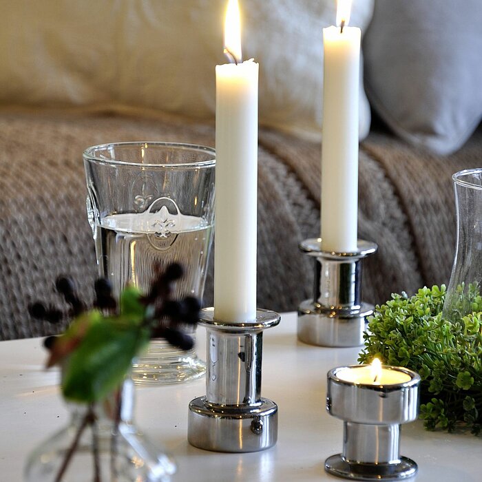 Drei brennende, weiße Müller Kerzen in Spiegelhalterungen, angeordnet auf einem Tisch zusammen mit einem Glas Wasser, einer kleinen Vase mit dunklen Beeren und grünen Blättern, sowie einem grünen Pflanzenarrangement. Im Hintergrund ist ein grau-beiges Sof