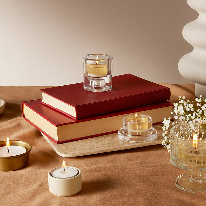 Vier brennende Teelichter von Bolsius in gläsernen und metallenen Teelichtern. Die Kerzen stehen auf einem braunen Tuch, umgeben von zwei roten Büchern, einer weißen Vase mit weißen Blumen und einem cremefarbenen Kerzenständer.