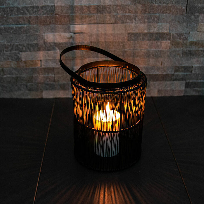 Eine brennende Kerze steht in einem schwarzen, drahtigen Laternenhalter mit einem Griff. Die Laterne wirft ein stimmungsvolles, warmes Lichtmuster auf die umgebende Fläche. Im Hintergrund ist eine Wand mit einer rustikalen Steinverkleidung zu sehen.
