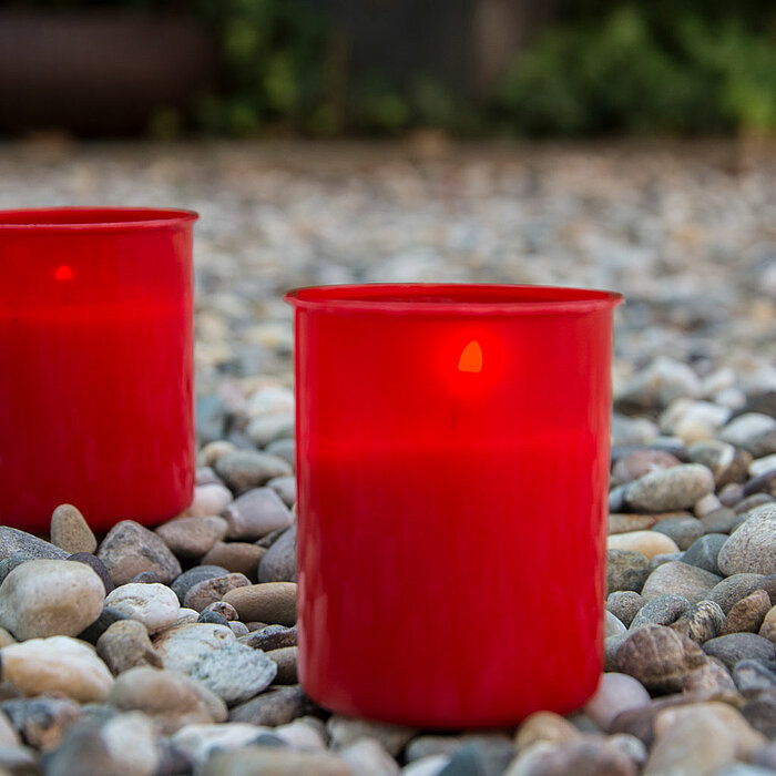 Zwei brennende rote Bolsius Grabkerzen stehen auf einem Bett aus kleinen, glatt polierten Kieseln im Freien. Die Kerzen strahlen in der Dämmerung ein warmes Licht aus und scheinen eine ruhige, besinnliche Atmosphäre zu schaffen. Die natürliche Kulisse aus