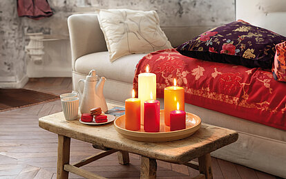 Brennende Stumpenkerzen in verschiedenen Größen, auf einem rustikalen Tisch in einem gemütlichen Wohnzimmer. Die Stumpenkerzen sind in Rot-, Orange, und Elfenbeintönen fabrlich aufeinander abgestimmt.