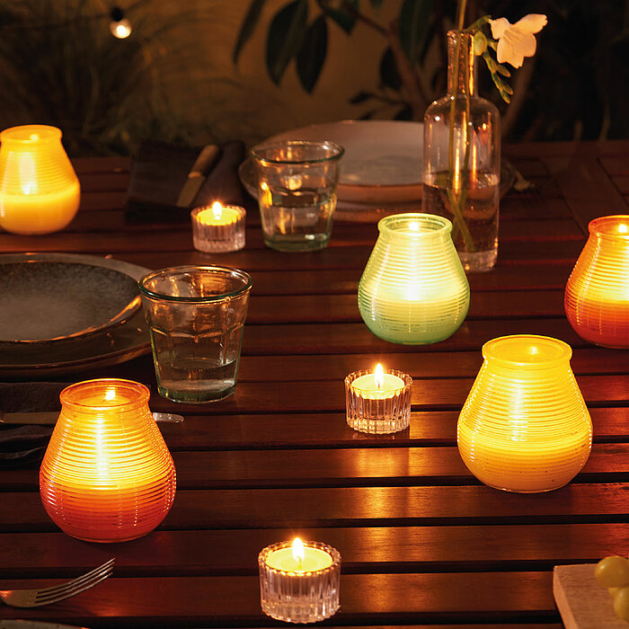 Bolsius Windlichter in verschiedenen Farben, darunter Gelb, Türkis und Orange, stehen auf einem Holztisch neben brennenden Teelichtern. Der Tisch ist mit Glasgeschirr, einem Teller, Besteck und einer kleinen Vase mit einer einzelnen weißen Blume dekoriert
