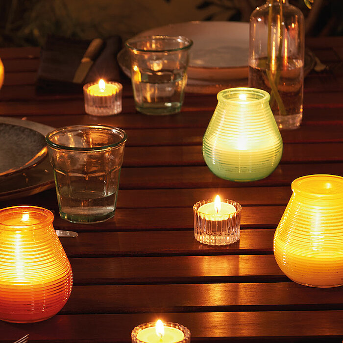 Bolsius Teelichter in transparenten Glasbehältern und bunten Kerzenhaltern stehen auf einem Holztisch. Die warmen Flammen der Teelichter schaffen eine gemütliche Atmosphäre. Um die Teelichter herum sind Wassergläser, Teller und Besteck arrangiert. Eine Va