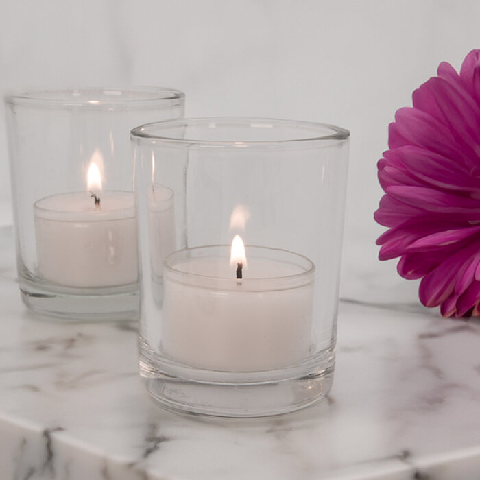 Zwei brennende Teelichter in klaren Glasbehältern auf einer eleganten Marmorplatte. Daneben liegt eine einzelne, kräftig pinkfarbene Blume. Die sanfte Beleuchtung und der weiße Hintergrund verleihen der Szene eine ruhige und friedliche Stimmung.