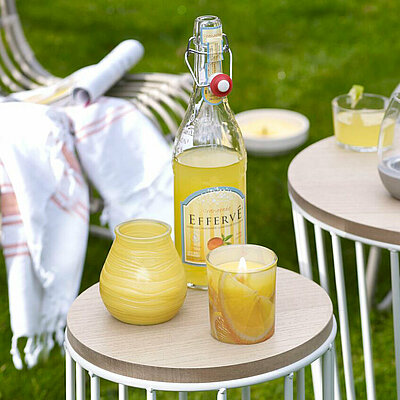 Brennende Citronella Duftkerzen stehen auf zwei Beistelltischen vor Flechtstühlen im Garten. Zwei Patio Windlichter, ein Glas mit Zitronenmotiv und ein Beton-Kerzenhalter mit einem Teelicht darin. Alle Kerzen sind gelb, genau wie die französische Limonade in einer Bügelflasche, die neben den Gläsern auf dem Tisch steht.