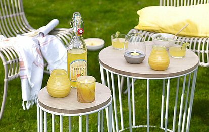 Brennende Citronella Duftkerzen stehen auf zwei Beistelltischen vor Flechtstühlen im Garten. Zwei Patio Windlichter, ein Glas mit Zitronenmotiv und ein Beton-Kerzenhalter mit einem Teelicht darin. Alle Kerzen sind gelb, genau wie die französische Limonade in einer Bügelflasche, die neben den Gläsern auf dem Tisch steht.