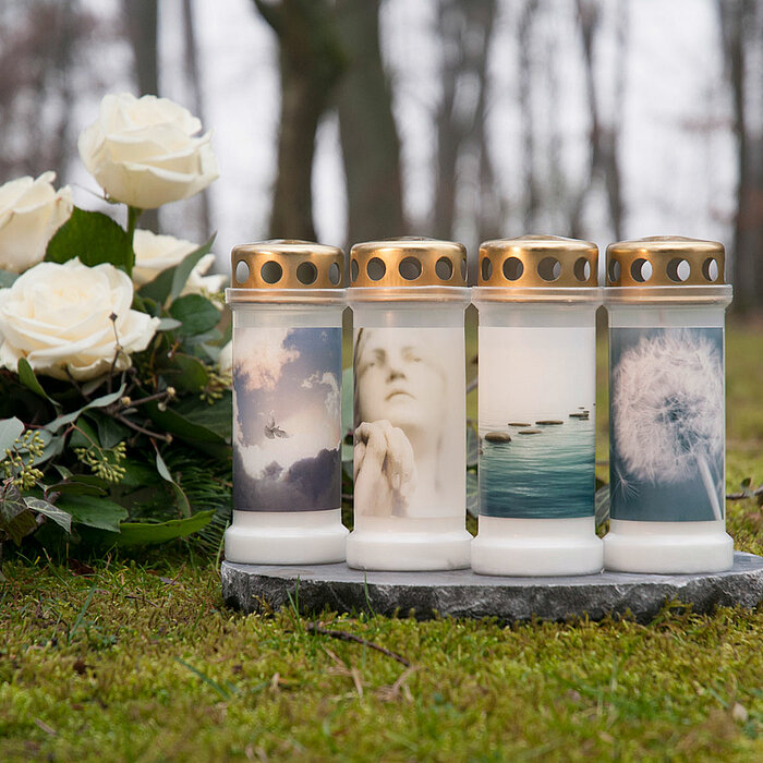 Vier Bolsius Grabkerzen in weißen Kunststoffgehäusen und mit goldenen Deckeln stehen nebeneinander auf einem grauen Stein in einer grasigen Umgebung. Die Grabkerzen sind jeweils mit verschiedenen Bildern verziert, wie Wolken, einem Gesicht und einer Puste