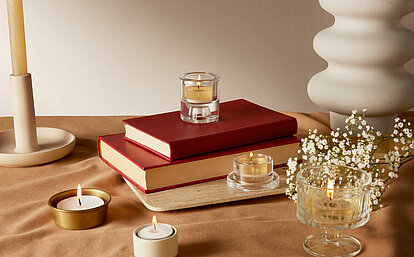 Das Bild ist in cremefarbenen und braunen Tönen gehalten. Auf einer beigefarbenen Decke liegen drei rot eingebundenen Bücher und unterschiedliche Teelichthalter mit verschieden großen weissen brennenden Teelichtern. Am linken Bildrand sieht man einen cremefarbenen Stabkerzenhalter. Am rechten Bildrand sieht man eine weisse bauchige Vase.