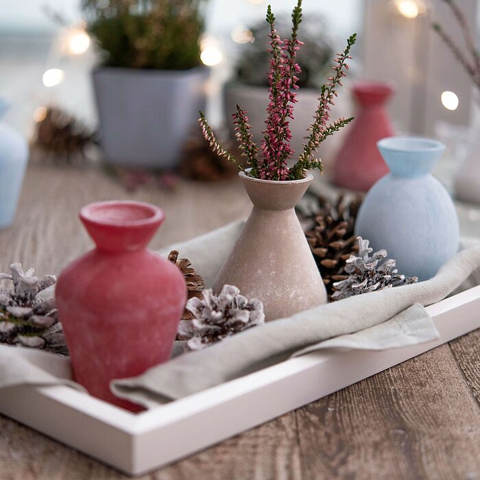 Drei kleine Vasen in den Farben Rot, Beige und Blau stehen auf einem weißen Tablett, das mit einem hellgrauen Tuch ausgelegt ist. Neben den Vasen befinden sich dekorative Elemente wie Tannenzapfen und frostige Zweige. Im Hintergrund sind weitere Vasen in 
