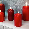 Fünf brennende rote Stumpenkerzen in verschiedenen Größen stehen auf einer weißen Oberfläche vor einem dezent gemusterten grauen Hintergrund. Die Produkte sind von DecoLite.