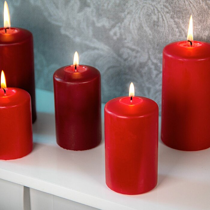 Fünf brennende rote Stumpenkerzen in verschiedenen Größen stehen auf einer weißen Oberfläche vor einem dezent gemusterten grauen Hintergrund. Die Produkte sind von DecoLite.