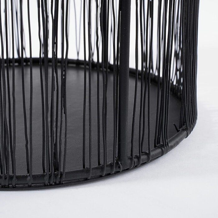 Ein detailreiches Bild zeigt den unteren Teil eines schwarzen, runden Drahtkorbs, der auf einer weißen Fläche steht. Die dünnen, vertikalen Drahtstreben des Korbs sind ungleichmäßig verteilt und verleihen dem Gegenstand ein modernes und minimalistisches A