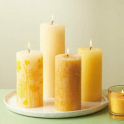 Ein Arrangement aus Frühlingsdeko auf einem hellgrünen Tisch. Auf einem weißen Tablett stehen brennende Rustikkerzen in verschiedenen Gelbtönen, darunter auch Kerzen mit fröhlichem Blumenmotiv. Daneben steht ein brennendes Dutkerzenglas mit gelbem Wachs, ein Raumduftdiffuser mit dekorativen Rattanstäbchen und ein brauner Tonkrug, in dem ein Zweig mit gelben Blüten platziert wurde.