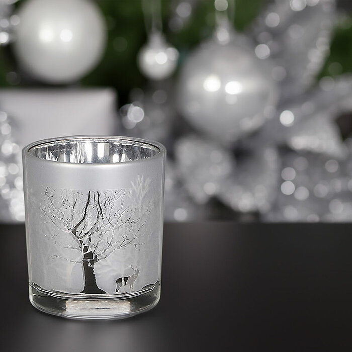 Ein einziges Teelichtglas mit einem Baum- und Hirschmotiv in mattem Weiß von Edelman, steht auf einer dunklen Oberfläche. Im Hintergrund funkelt weihnachtliche Dekoration in Silber mit reflektierenden Kugeln und Zweigen. Die Atmosphäre ist festlich und el
