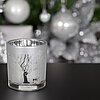 Ein einziges Teelichtglas mit einem Baum- und Hirschmotiv in mattem Weiß von Edelman, steht auf einer dunklen Oberfläche. Im Hintergrund funkelt weihnachtliche Dekoration in Silber mit reflektierenden Kugeln und Zweigen. Die Atmosphäre ist festlich und el
