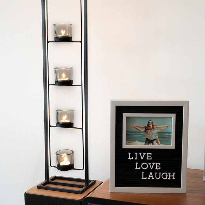 Vier brennende Kerzen in kleinen Gläsern sind in einem stilvollen, schwarzen Metall-Kerzenhalter vertikal angeordnet. Daneben steht ein eingerahmtes Bild mit dem Zitat "LIVE LOVE LAUGH" und einem Foto von einer Person im Wasser mit ausgestreckten Armen. B