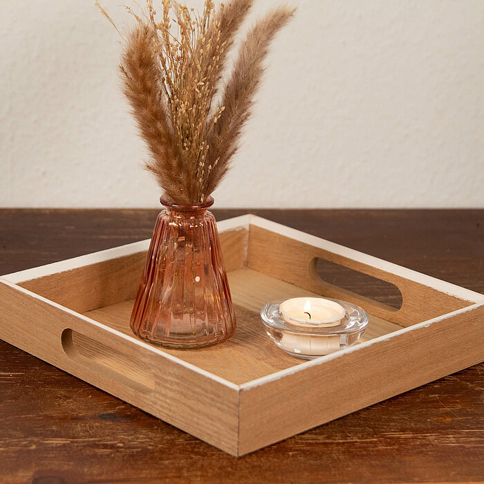 Eine brennende Teelichtkerze in einem durchsichtigen Kerzenhalter und eine Vase mit getrockneten Gräsern stehen auf einem hölzernen Tablett mit Tragegriffen. Das Tablett befindet sich auf einem rustikalen Holztisch. Die Szene wirkt gemütlich und schlicht.