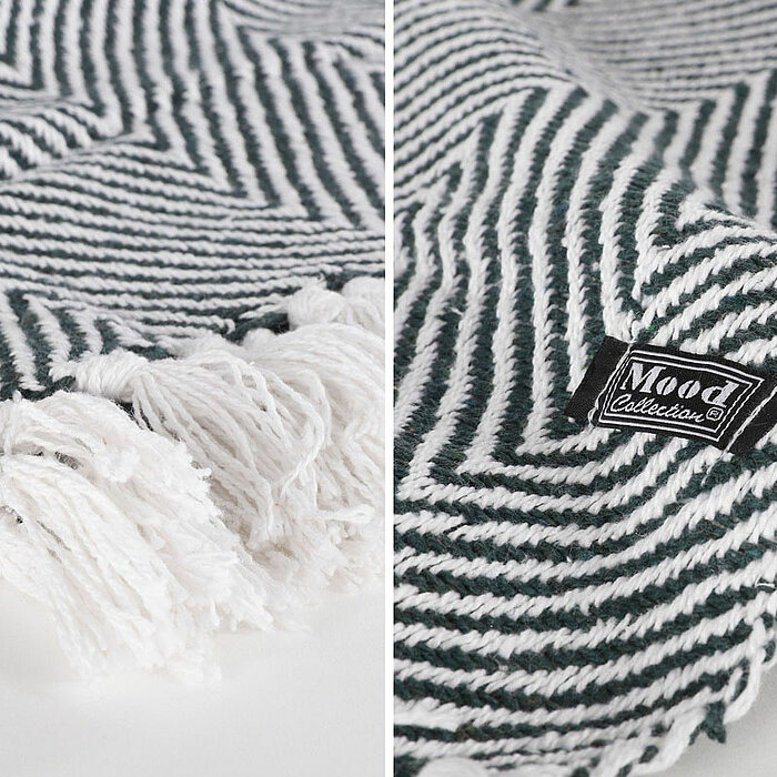 Das Bild zeigt zwei grüne Zimmerpflanzen in strukturierten, braunen Pflanzgefäßen, die auf einem Holzboden stehen. Im Hintergrund liegen zusammengerollte graue Decken in einem geflochtenen Korb, der neben einem gelben Kissen mit grauer Umrandung steht. An