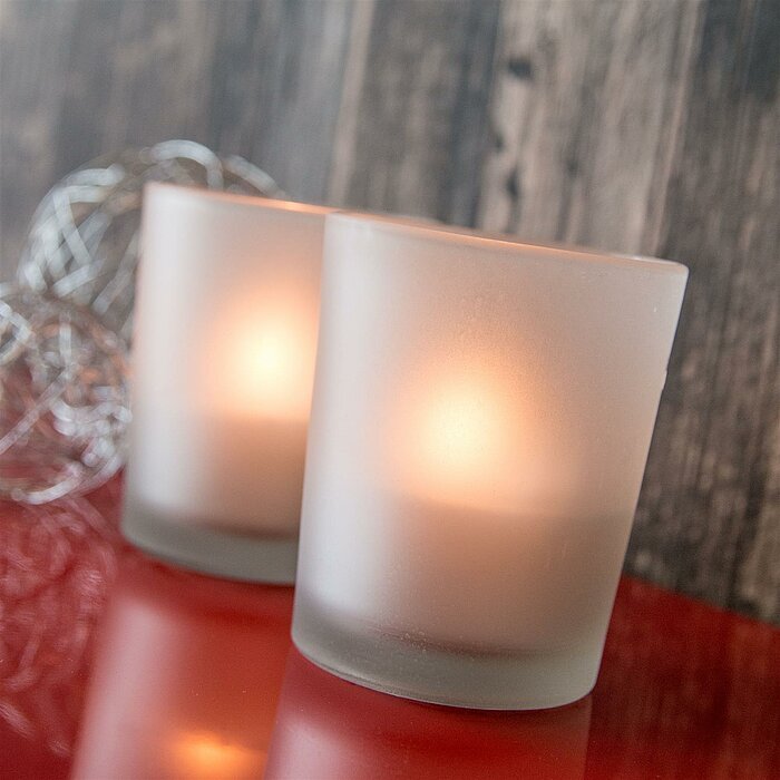 Vier brennende Sandra Rich Teelichter in matt-weißen Glashaltern stehen in einer geraden Linie auf einem hellen Tisch. Die Hintergrundbeleuchtung erzeugt ein warmes, gemütliches Licht, das durch die matte Oberfläche der Gläser gedämpft wird. Ein grauer, u