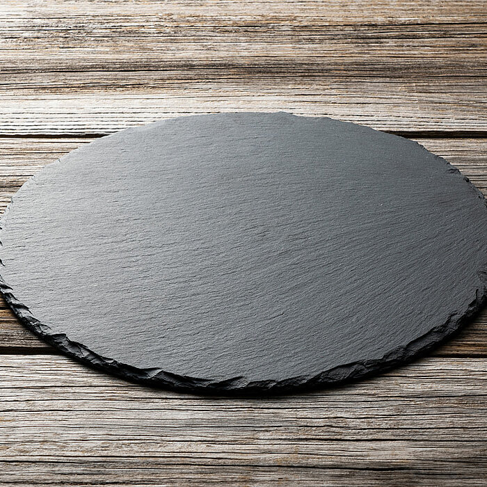 Eine runde, schwarze Schieferplatte liegt auf einem Tisch aus verwittertem Holz mit sichtbaren Maserungen. Die Platte hat unregelmäßig gearbeitete Kanten und eine natürliche, matte Oberfläche. Die rustikale Holzstruktur des Tisches bildet einen schönen Ko