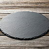 Eine runde, schwarze Schieferplatte liegt auf einem Tisch aus verwittertem Holz mit sichtbaren Maserungen. Die Platte hat unregelmäßig gearbeitete Kanten und eine natürliche, matte Oberfläche. Die rustikale Holzstruktur des Tisches bildet einen schönen Ko