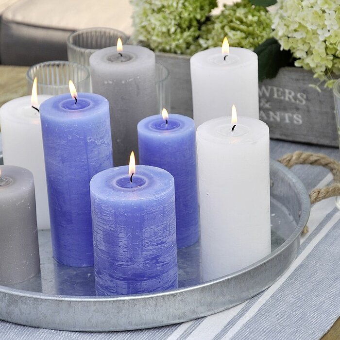 Acht brennende Müller Kerzen in verschiedenen Größen und Farben (Blau, Weiß und Grau) stehen auf einem runden Tablett aus Metall, das auf einem hölzernen Tisch platziert ist. Neben dem Tablett stehen Wassergläser und eine Kiste mit weißen Blumen. Im Hinte