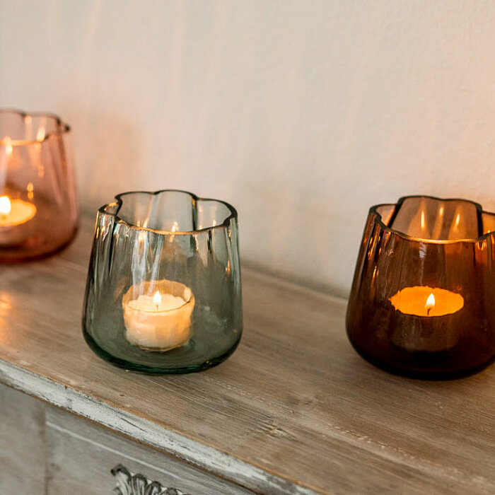 Drei Teelichthalter aus gefärbtem Glas vom Hersteller Sandra Rich in den Farben Rosa, Grün und Braun stehen auf einer Holzkommode. Sie enthalten jeweils ein brennendes Teelicht. Daneben steht eine große cremefarbene Stumpenkerze, ebenfalls brennend. Die s