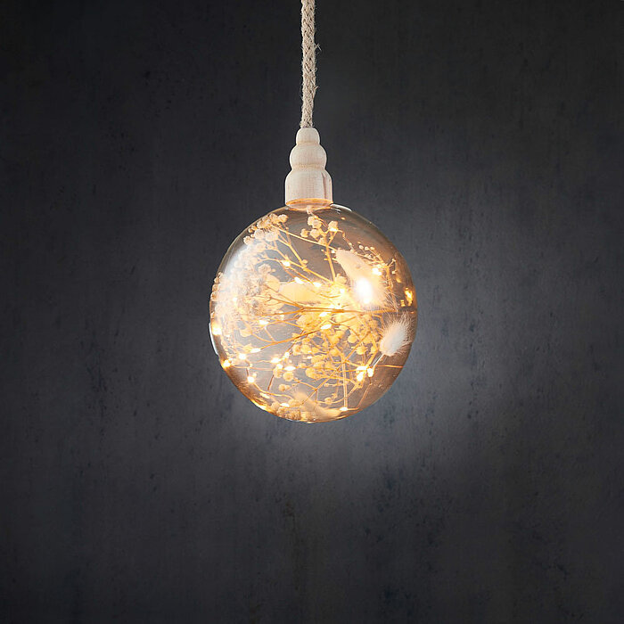 Eine runde, transparente Glühbirne mit einem Gewinde aus hellem Holz hängt vor einem dunklen Hintergrund. Innerhalb der Glühbirne sind zarte Pflanzen- und Blumenmuster sowie kleine Lichter sichtbar, die ein warmes, diffuses Licht abstrahlen und eine gemüt