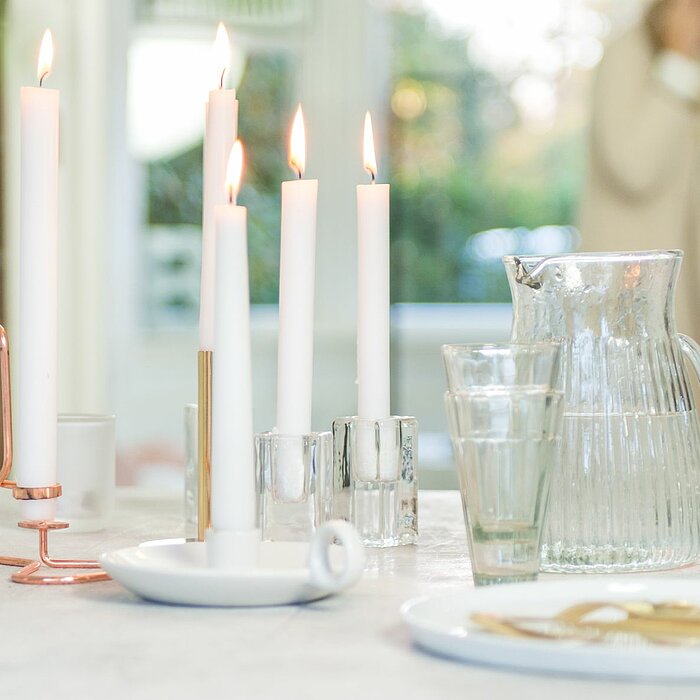 Fünf brennende Bolsius Tafelkerzen in Weiß stehen auf einem Tisch, in verschiedenen Haltern aus Glas und einem modernen Kerzenhalter aus Kupfer. Auf dem Tisch befinden sich zudem eine Glas-Karaffe, zwei leere Trinkgläser und ein Teller mit goldfarbenem Be