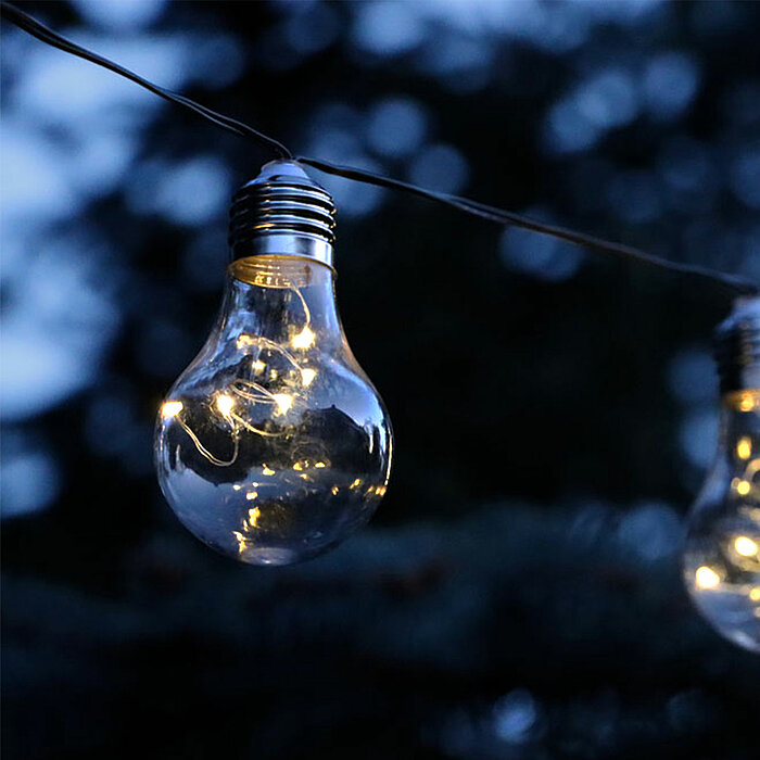 Eine Reihe von Glühbirnen ist an einem Draht aufgehängt und leuchtet in der Abenddämmerung. Die Lichter strahlen warmes, gemütliches Licht aus. Im Hintergrund sind unscharf dunkle Bäume und der Abendhimmel zu erkennen, was eine ruhige und friedliche Atmos