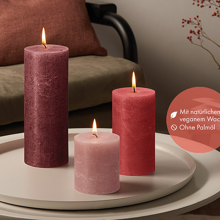 Sechs brennende Bolsius Rustikkerzen in verschiedenen Größen und Farben, darunter Weiß, Grün und Rosa, stehen auf einer Holzablage. Daneben befinden sich eine rosa Blume in einer kleinen Vase und ein Set aus Keramikgeschirr. Die Szene wirkt warm und einla