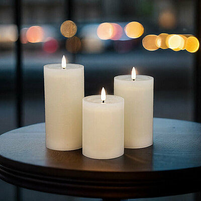 Drei leuchtende LED Stumpenkerzen in verschiedenen Größen stehen auf einem kleinen runden Holztisch. Im Hintergrund erkennt man durch ein großes Schaufenster eine Pariser Straße in der Abenddämmerung.