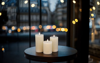 Drei leuchtende LED Stumpenkerzen in verschiedenen Größen stehen auf einem kleinen runden Holztisch. Im Hintergrund erkennt man durch ein großes Schaufenster eine Pariser Straße in der Abenddämmerung.