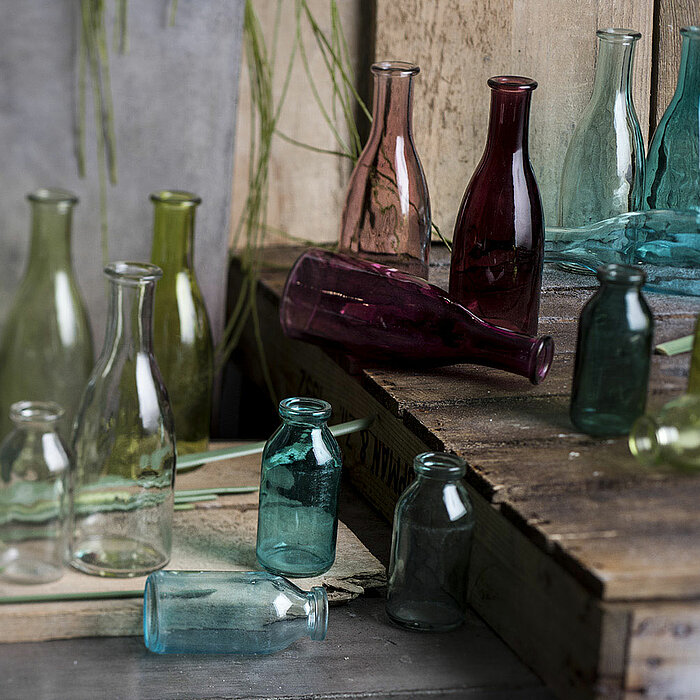 Zwei Glasvasen vom Hersteller "Sandra Rich" in den Farben Grün und Orange stehen auf einer rustikalen Holzoberfläche mit Moos. Neben ihnen befindet sich eine weitere, teilweise sichtbare Vase in Blau.