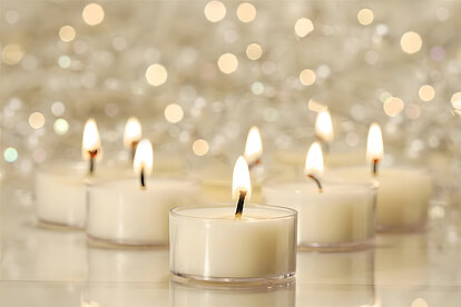 In der Mitte des Bildes sind in der Frontalansicht acht brennende Teelichter im klaren Becher von Wenzel zu sehen. Ganz vorne steht ein einzelnes Teelicht, während die restlichen sieben Teelichter dahinter in einer geraden Linie angeordnet sind. Der Hintergrund zeigt einen hell erleuchteten Raum, der in weißen und cremefarbenen Tönen gehalten ist. Die Flammen der Teelichter und die harmonischen Farben des Raums schaffen eine ruhige und etwas festliche Atmosphäre.