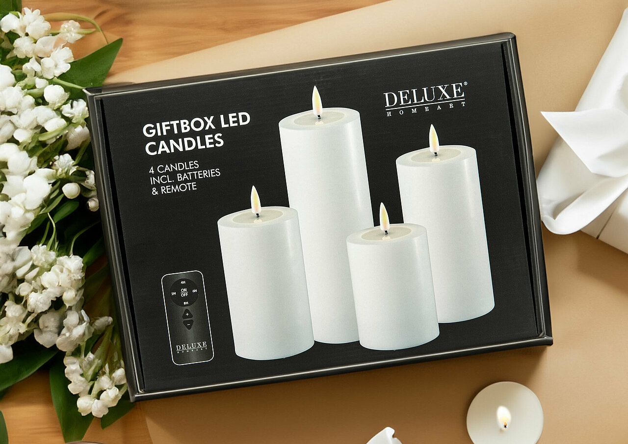 Von oben sieht man auf einen hellbraunen Tisch, auf dem einige brennende Teelichter und Kerzen im Glas verteilt sind. In der Mitte des Bildes befindet sich eine LED-Kerzen-Geschenkbox von Deluxe Homeart. Links neben der Box liegt ein Strauß Maiglöckchen.