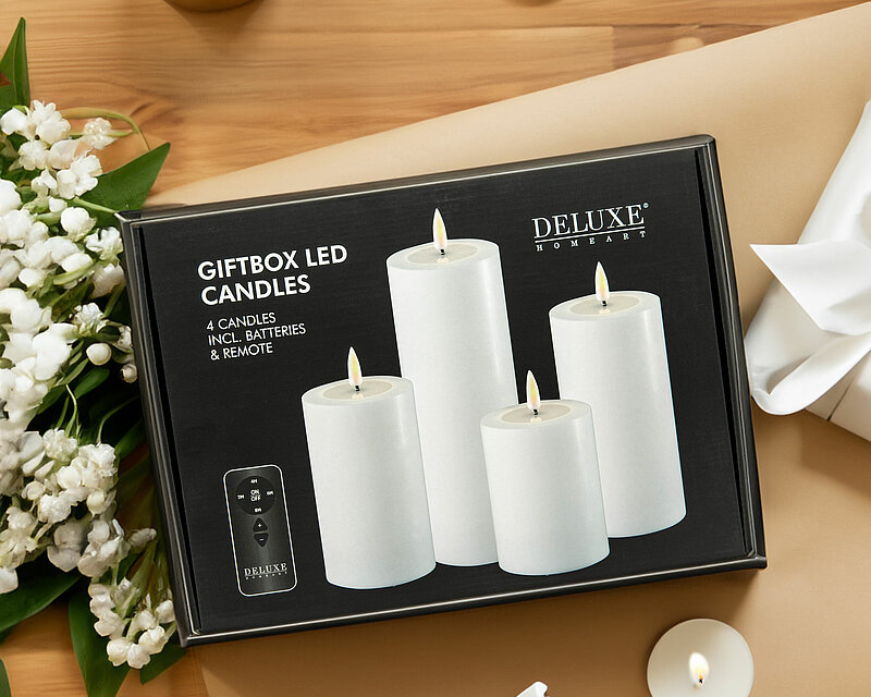 Von oben sieht man auf einen hellbraunen Tisch, auf dem einige brennende Teelichter und Kerzen im Glas verteilt sind. In der Mitte des Bildes befindet sich eine LED-Kerzen-Geschenkbox von Deluxe Homeart. Links neben der Box liegt ein Strauß Maiglöckchen.