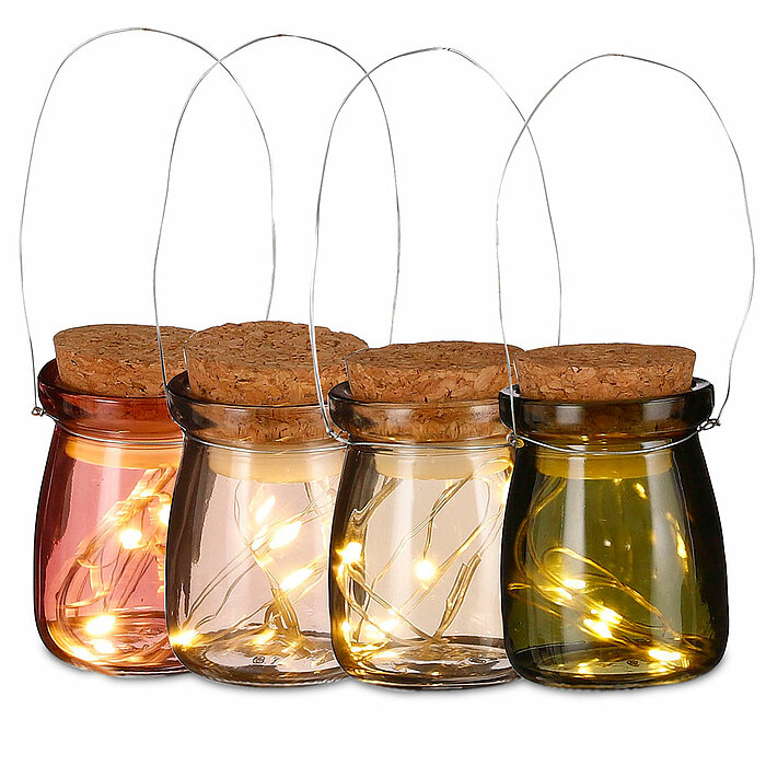 Vier Glaslaternen mit Korkdeckeln und Drahtgriffen, jede gefüllt mit kleinen, warmweiß leuchtenden LED-Lichterketten. Die Laternen haben unterschiedliche Farben: Rosa, Hellbraun, Klar und Grün. Die stimmungsvolle Beleuchtung der Lichterketten schafft eine