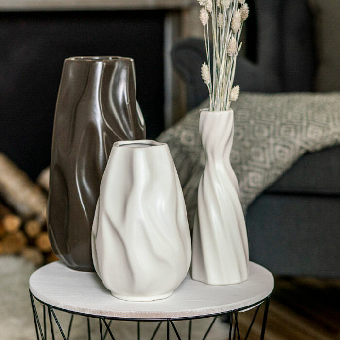 Acht Vasen vom Hersteller "Sandra Rich" stehen auf einem Tisch, der mit einer weißen Tischdecke und einem Leinentuch dekoriert ist. Die Vasen sind in Pastelltönen gehalten, darunter verschiedene Nuancen von Blau und Rosa. Die Vasen haben unterschiedliche 