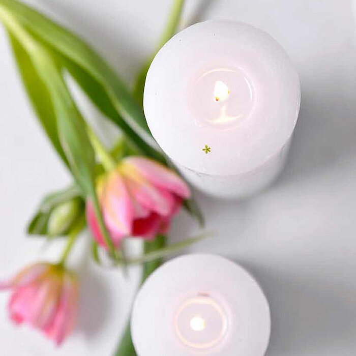 Vier brennende gelbe Kerzen von Müller in verschiedenen Größen stehen auf einem rechteckigen weißen Kerzenhalter auf einem hellen Tisch. Im Hintergrund befindet sich eine graue Vase mit einem bunten Blumenstrauß und eine weiße dekorative Skulptur.