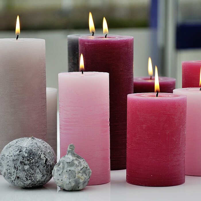 Acht brennende Müller Stumpenkerzen in verschiedenen Größen und Farben, darunter Rosa, Pink, Rot und Grau, stehen auf einem weißen Tisch. Vor den Kerzen liegen zwei Dekosteine.